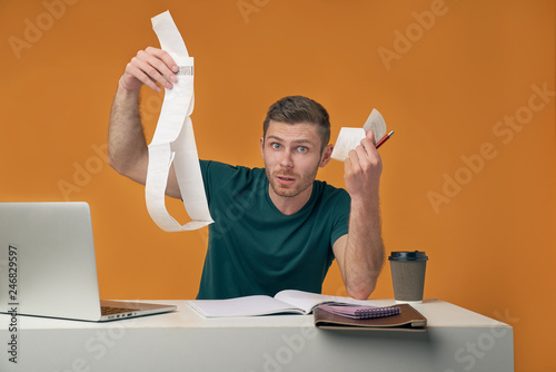 The indignant man holds checks, not understand from where the big sums, not ready to unforeseen expenses. Isolated on an orange background in the Studio photo