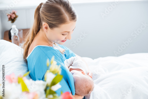 side view of happy young mother breastfeeding newborn baby on bed in hospital room