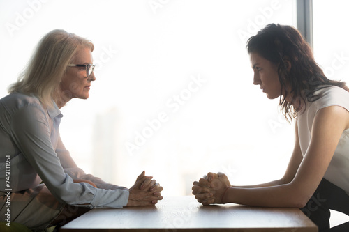 Young and old businesswomen with clasped hands looking at each other sitting opposite as rivalry confrontation concept, female career success envy and jealousy, generations conflict at work concept photo