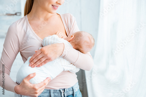 cropped shot of smiling young mother standing and breastfeeding infant baby at home photo