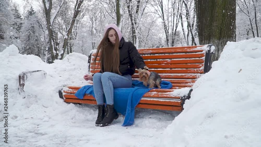 Young woman with long hair covered with a hood smoking on bench in a winter snow-covered park. Teenager and a yorkie resting outdoors together. Slow motion.