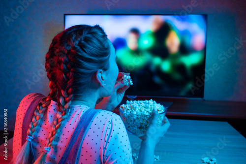 A young girl watching movies and eating popcorn with a bowl on the background of the TV. The color bright lighting, blue and red. Relax, rest at home when watching TV, film. Background for design