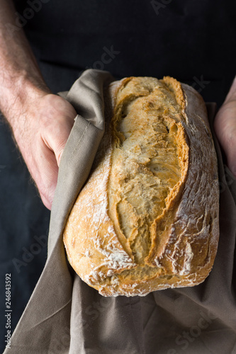 Male chef baker holds in hands in grey cotton towel freshly baked homemade loaf of bread. Authentic style. Lifestyle photo. Mediterranean French cuisine panification