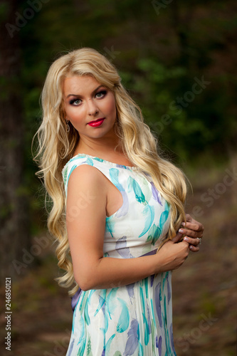 Young beautiful blonde woman in white dress with blue and green flowers posing in forest in summer