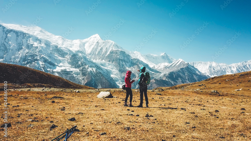 A couple walking on the Annapurna Circuit Trek, Himalayas, Nepal. Annapurna chain in the back, covered with snow. Clear weather, dry grass, snowy peaks. High altitude, man pointing to the peaks.