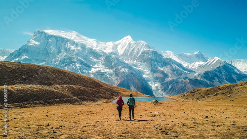 A couple walking towards the Ice lake, as part of the Annapurna Circuit Trek, Himalayas, Nepal. Annapurna chain in the back, covered with snow. Clear weather, dry grass, snowy peaks. High altitude photo