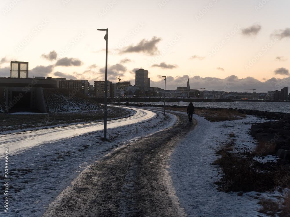 Snow-covered street in Reykjavik city. Reykjavik is the capital of Iceland. Photographed in October, in the evening. There's a person walking. In the background, there are buildings.