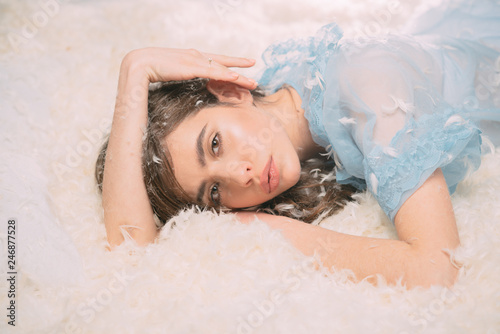 I want to stay in bed. Pretty girl in nightie lingerie with feather in long hair. Cute girl relaxing on feather bed pillow and mattress. Young lady wear silky blue nightie. Making up the bed to sleep