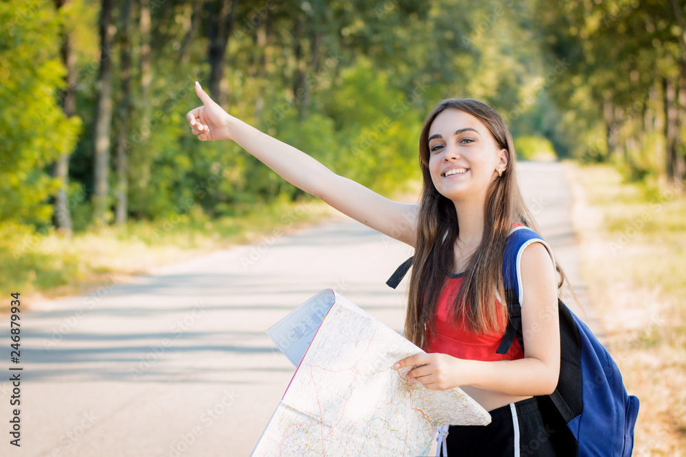 Naklejka premium Smiling girl traveling hitchhiking putting up a hand tries to stop the car. Traveler girl with heavy backpack and a map in her hands wants to stop a car to get a lift to a destination