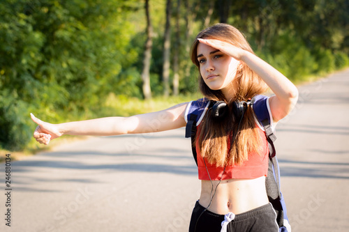 Exhausted girl standing at the scorching summer sun looking away while keeping his hand over her eyes tries to stop an approaching car at the road in the countryside tired of traveling in a hot day