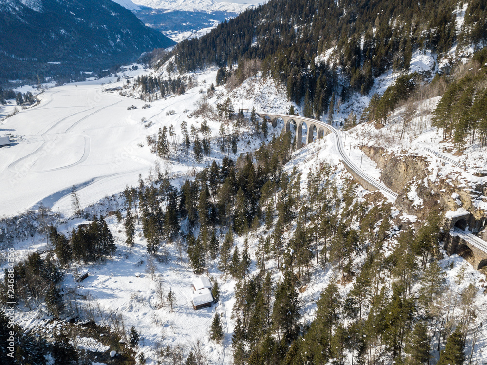 Aerial image of a mountain valley in Alps in snowy winter season with railway viaduct at the background
