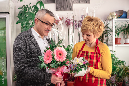 Husband buying flowers and gifts for Valentine's day for his wife in local flower shop, saleswoman showing him some bouquets