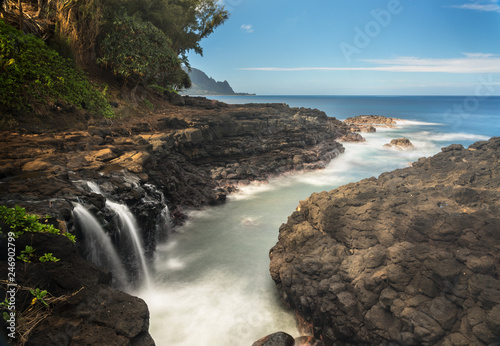 Rugged rocks around small waterfall falling into the ocean with the Na Pali coastline by Hanalei in the distance