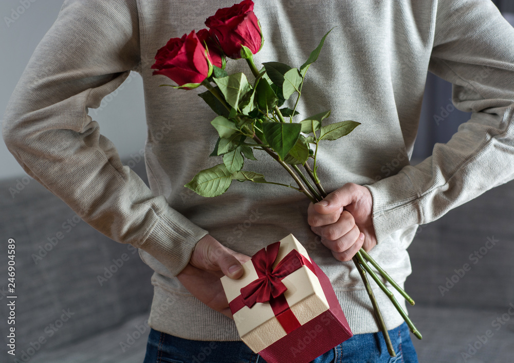 Man holding red rose flowers and gift box behind his back at home.  Surprised gift for woman. Romantic and valentines day holiday concept.  Close up, selective focus Stock Photo | Adobe Stock