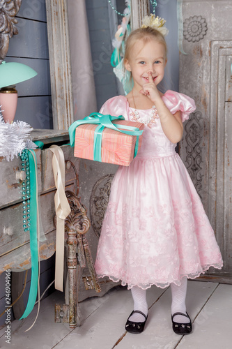 Portrait of cute european little blonde princess girl with crown in beautiful dress in decorated studio in christmas location with toys and christmas tree