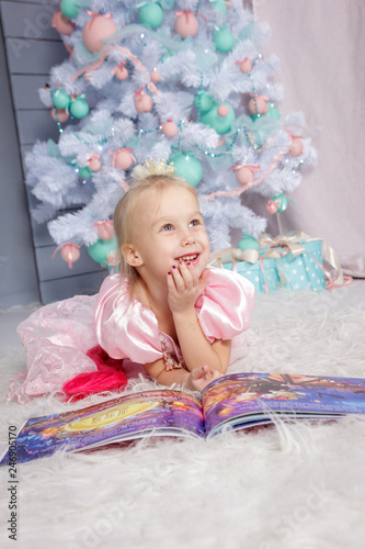 Portrait of cute european little blonde princess girl with crown in beautiful dress lying on the floor and reading book in decorated studio in christmas location with toys and christmas tree