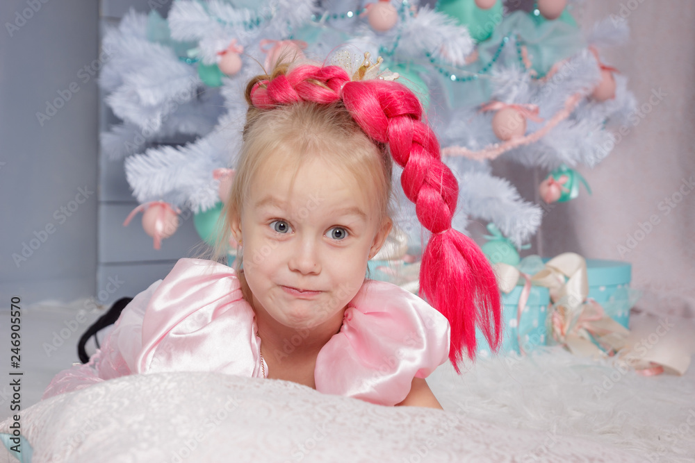 Fototapeta premium Portrait of cute european little blonde princess girl with crown in beautiful dress in decorated studio in christmas location with toys and christmas tree