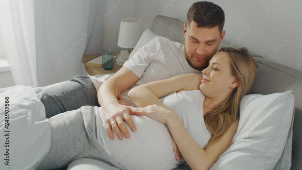 Happy Young Couple Cuddling Together in the Bed, Young Woman is Pregnant and Loving Husband Holds Her In Embrace and Caresses Her Belly Tenderly Happy Young Couple Cuddling Together in the Bed, Young Woman is Pregnant and Loving Husband Holds Her In Embrace and Caresses Her Belly Tenderly
