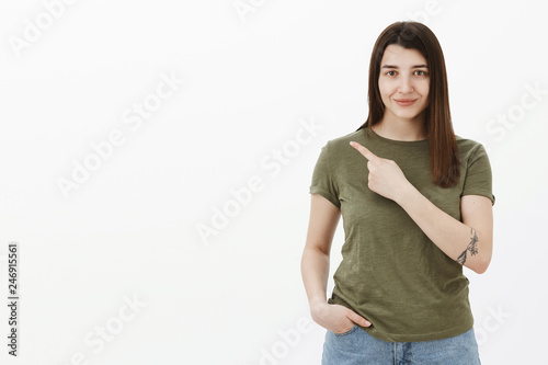 Look there. Portrait of confident and ambitious good-looking brunette in 20s with tattoo pointing at upper left corner with arm crossed over body and smiling pleased at camera as suggesting product © Cookie Studio