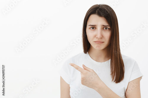 Gloomy sad and distressed cute brunette girl in white t-shirt frowning, pursing lips and pointing left with jealousy or regret, telling about unfair thing happened disappointed and upset © Cookie Studio