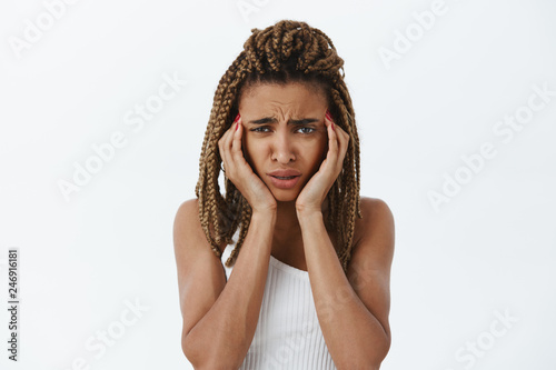 Waist-up shot of concerned and perplexed miserable dark-skinned female with dreadlocks frowning touching temples trying concentrate feeling headache or migraine over white background
