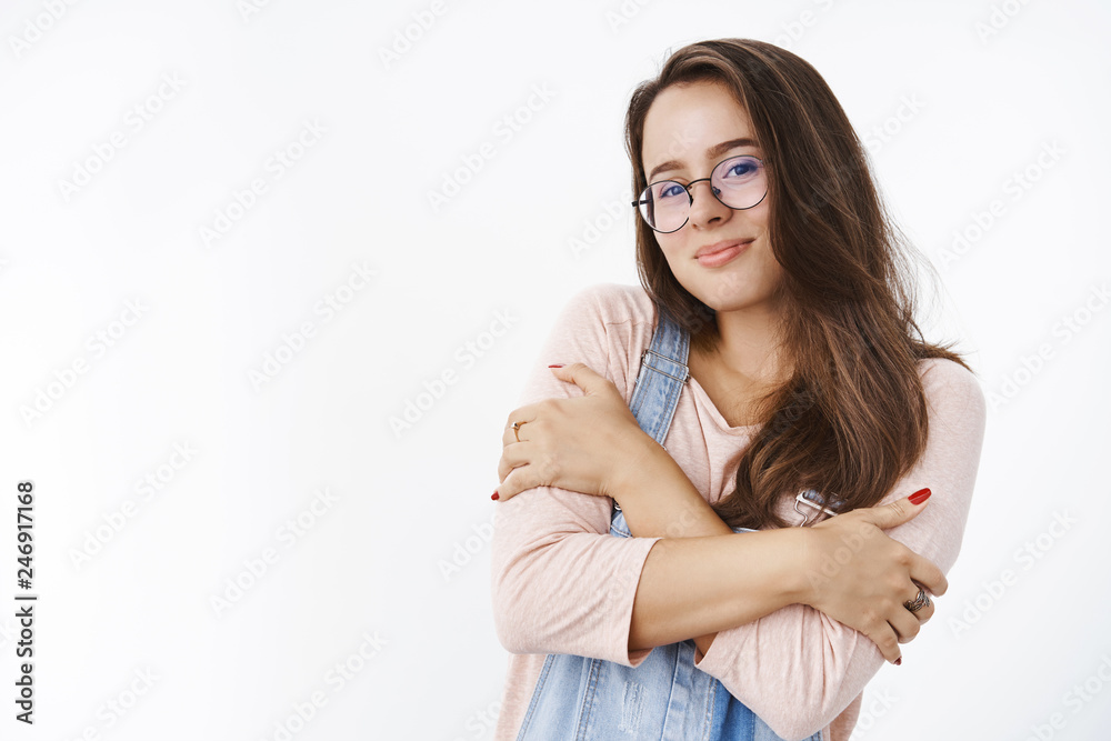 Studio shot of tender silly young brunette in glasses and overalls hugging herself with dreamy pretty smile as if feeling chilly, wanting cuddles in warm hands standing against gray background Studio shot of tender silly young brunette in glasses and overalls hugging herself with dreamy pretty smile as if feeling chilly, wanting cuddles in warm hands standing against gray background