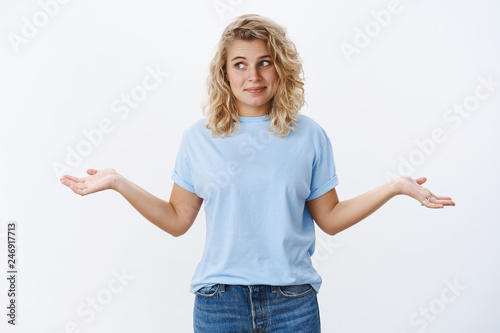 Who cares, I not know. Confused and unsure cute shrugging girl with blond curly hairstyle turning eyes left and smirking with hands spread sideways in unaware and clueless gesture over white wall