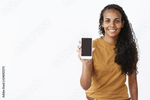 Waist-up shot of friendly creative and stylish attractive dark-skinned female holding smartphone showing cellphone screen at camera and smiling delighted as recommending device or app over gray wall