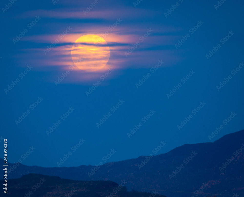 Full moon rising over a desert mountain in the Sonoran wilderness in southwest USA. The colorful full moon rises behind clouds in the night sky, giving the photograph texture in these photos 