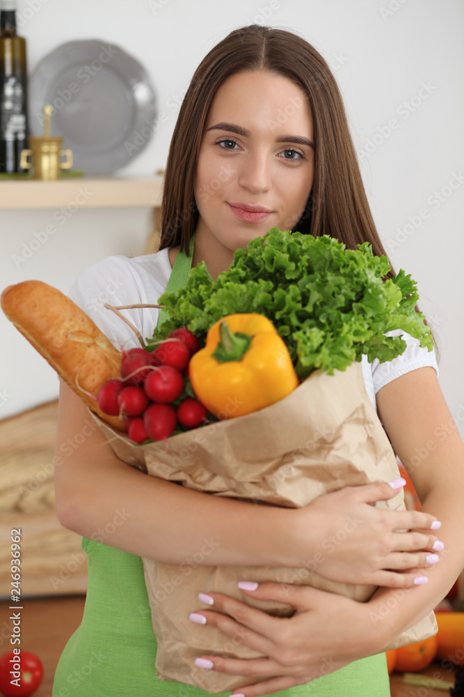 Young woman cooking in kitchen. Girl holding paper bag full of vegetables and fruits. Householding, tasty food and vegetarian in lifestyle concepts