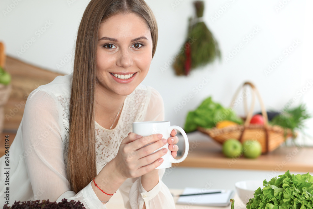 Young happy woman is holding white cup and looking at the camera while sitting at wooden table in the kitchen among green vegetables Young happy woman is holding white cup and looking at the camera while sitting at wooden table in the kitchen among green vegetables