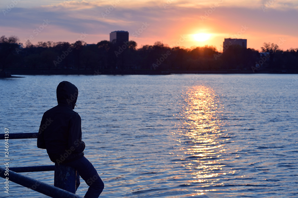 Single man watching sunset over a lake. Silhouette alone young man relax sitting on beach wooden walk way pier with sunset and cloud sky thinking Single man watching sunset over a lake. Silhouette alone young man relax sitting on beach wooden walk way pier with sunset and cloud sky thinking