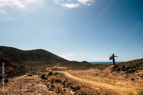 Happy hiker young woman with trekking backpack standing on stone in summer mountains - enjoying the desertic place with ocean in background - travel and freedom lifestyle concept