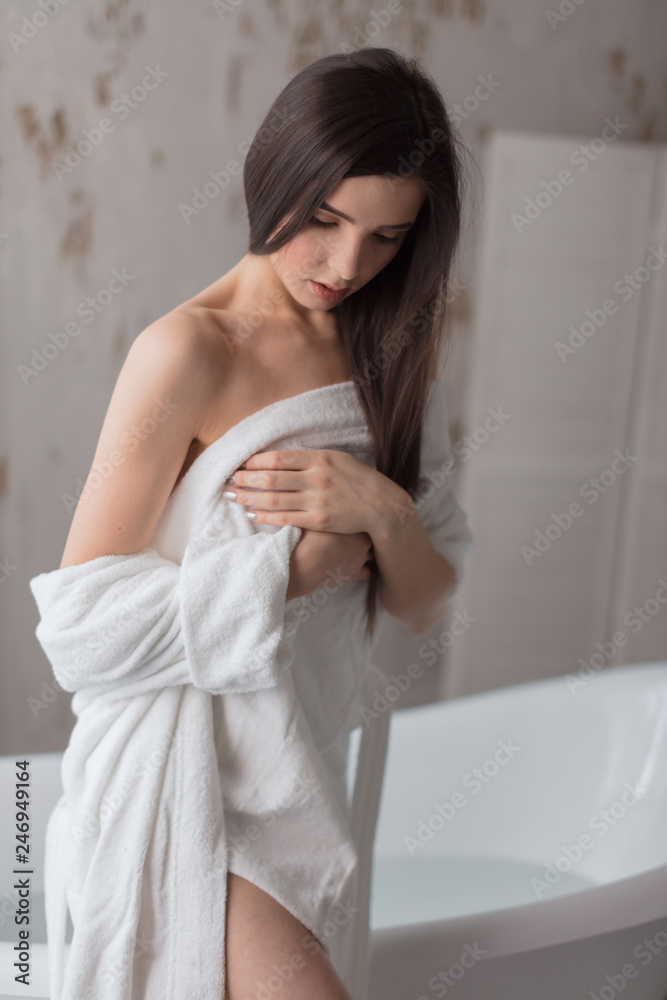 Young female model in a white bathrobe sitting at the side of freestanding white bath, bathroom design, luxury sanitary ware collection, preparing relaxing bath. Beauty, healthy lifestyle concept Young female model in a white bathrobe sitting at the side of freestanding white bath, bathroom design, luxury sanitary ware collection, preparing relaxing bath. Beauty, healthy lifestyle concept