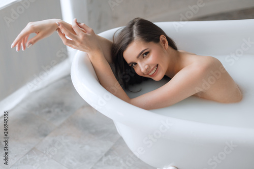 Happy satisfied naked girl with long wet brown hair smiles at camera while enjoying a bath with milk and hot steaming water. Spa body care.