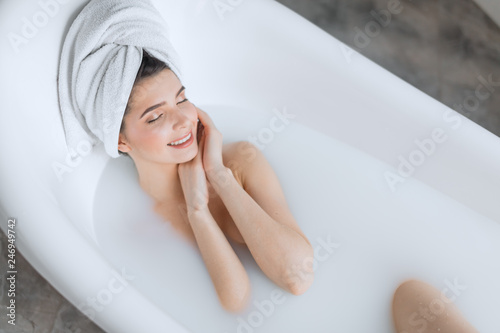 Gorgeous young brown-haired woman taking a relaxing milky bath, touching her clean fresh skin face and closes her eyes from bliss in a relaxing atmosphere.