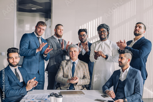 Diverse smiling business men gather at conference table together with his mature grey-haired leader, attending meeting or corporate briefing. African, indian and caucasian businessmen partners team.