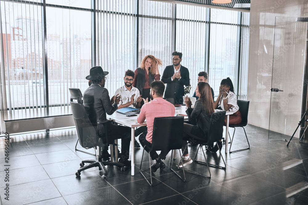 company is greeting foreign partner. happy diverse people are sitting at the table and clapping hands in the room with panorama window