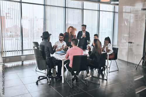 company is greeting foreign partner. happy diverse people are sitting at the table and clapping hands in the room with panorama window
