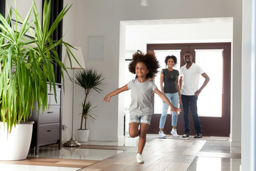Excited little funny african girl running exploring big modern house moving in, happy black parents and kid daughter coming into new home, cute mixed race child having fun in hallway, family mortgage