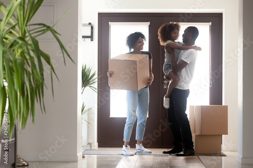 Happy black family with kid girl holding box entering into own house on moving day, african american parents and child standing in hallway, mortgage, relocation, tenants welcome to new home concept photo