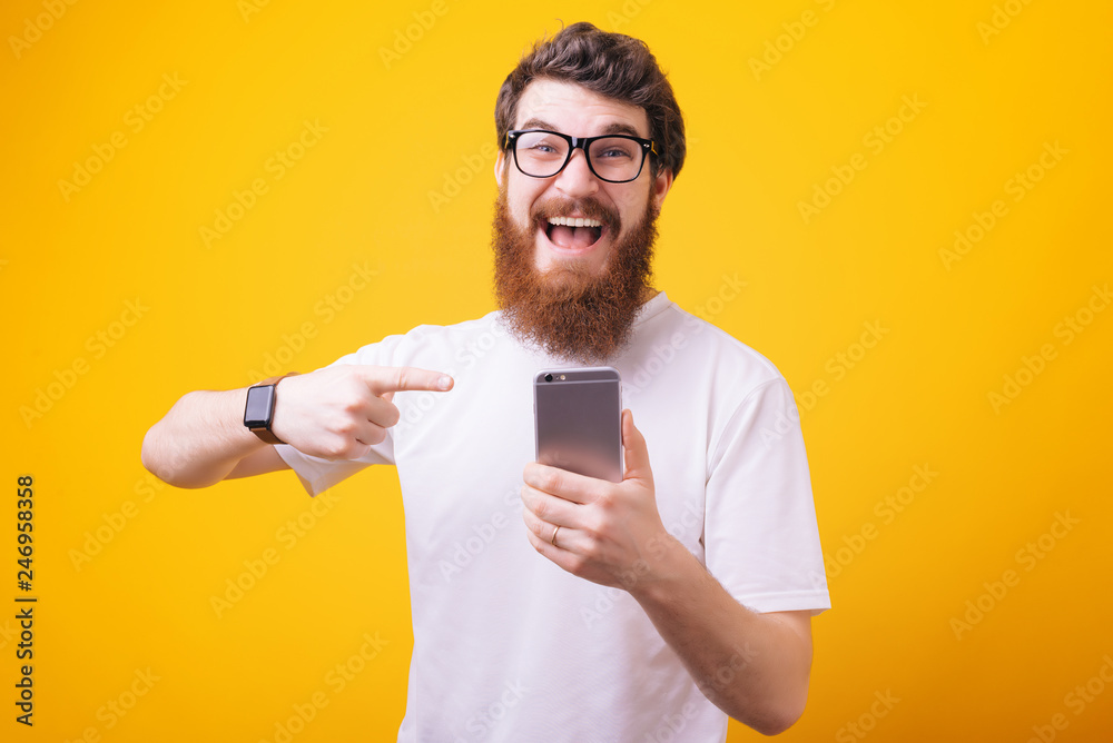 Promotion concept. Smiling young bearded man pointing at smartphone and looking at camera Promotion concept. Smiling young bearded man pointing at smartphone and looking at camera