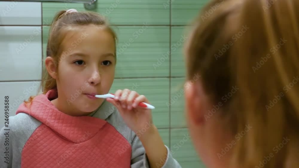 Close-up cute beautiful female child with fair hair brushes her teeth thoroughly in mirror reflection in bathroom on background wall from green and white tile Close-up cute beautiful female child with fair hair brushes her teeth thoroughly in mirror reflection in bathroom on background wall from green and white tile