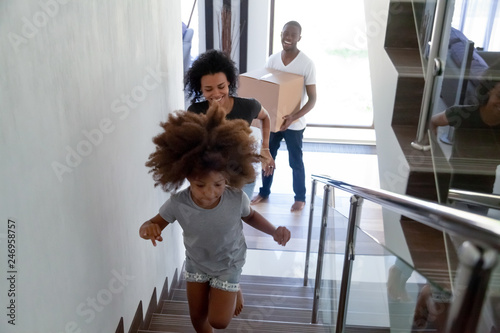 Happy cute black child running up stairs moving in two story modern house with parents carrying boxes  excited kid having fun going upstairs enjoying relocation into new home  african family mortgage