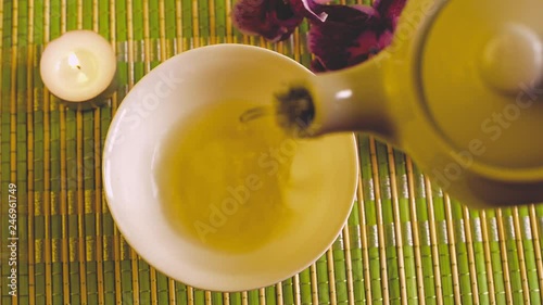 Green tea is poured into a small bowl that stands on a light mat next to a purple orchid flower and a burning candle. Top view.