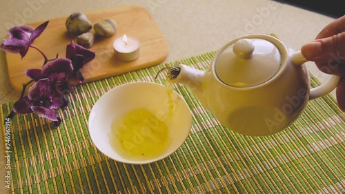 Green tea is poured into a small bowl that stands on a light table next to a purple orchid flower and a burning candle.
