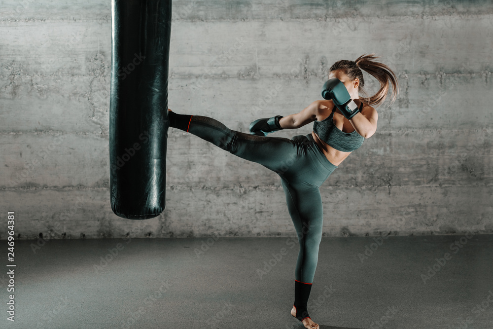 Caucasian woman in sportswear and with boxing gloves kicking bag in the  gym. Full length. Wall in background. Stock Photo | Adobe Stock