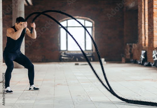 motivated man in stylish black sportswear tries to make a wave with a rope at gym, full length side view photo. copy space.motivation