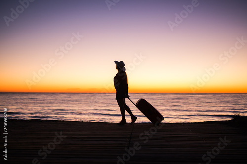 Traveling, beach and holiday concept - Female silhouette walking along ocean coast pulling suitcase