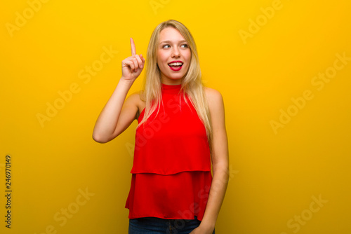 Young girl with red dress over yellow wall intending to realizes the solution while lifting a finger up